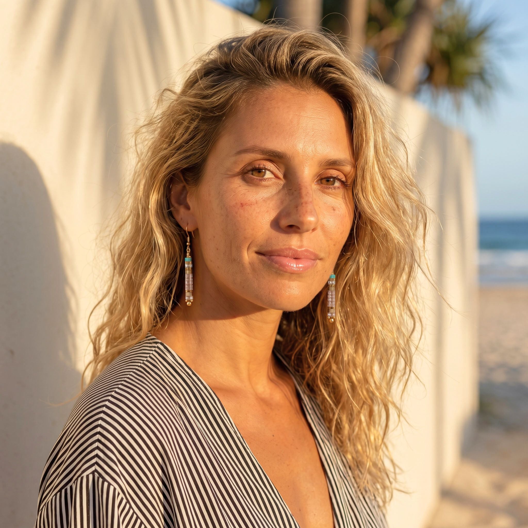 Woman with blonde hair and earrings standing on a beach with palm trees in the background