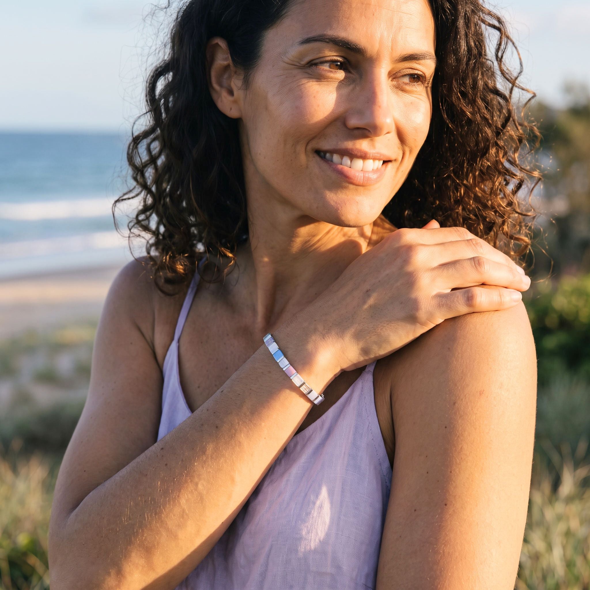 A smiling woman with curly brown hair stands outdoors near the beach, wearing a slim metallic beaded bracelet on her wrist.