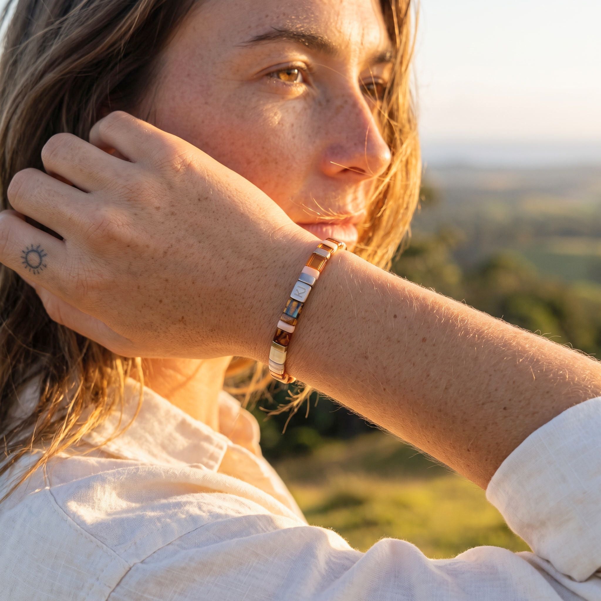 A close-up lifestyle shot of a woman wearing a slim metallic cuff bracelet with alternating rose gold and silver rectangular links, set against a blurred outdoor landscape background.