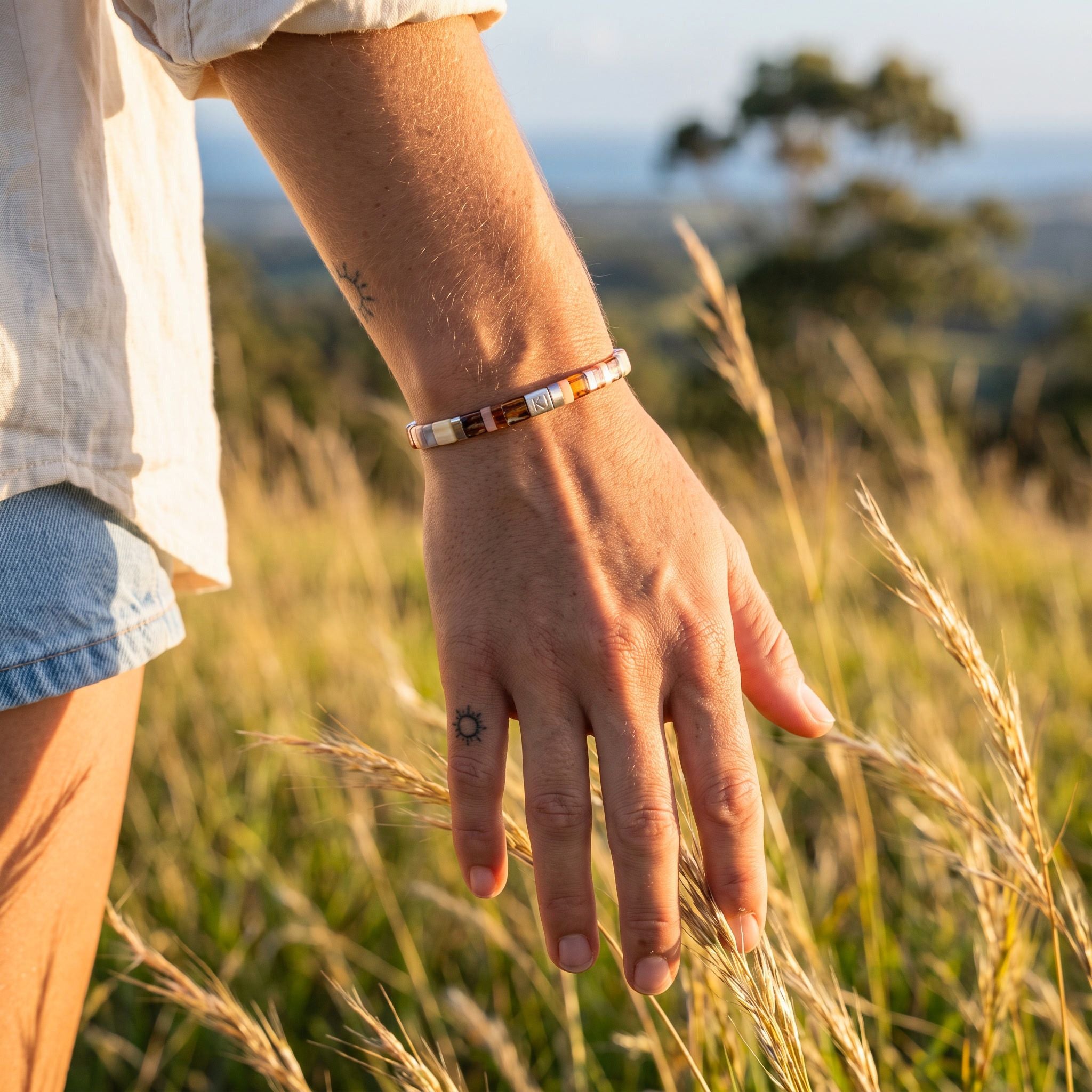 A person outdoors in a grassy field wearing a beaded bracelet on their wrist. The bracelet features a mix of earthy tones including brown, orange, cream, and white beads.
