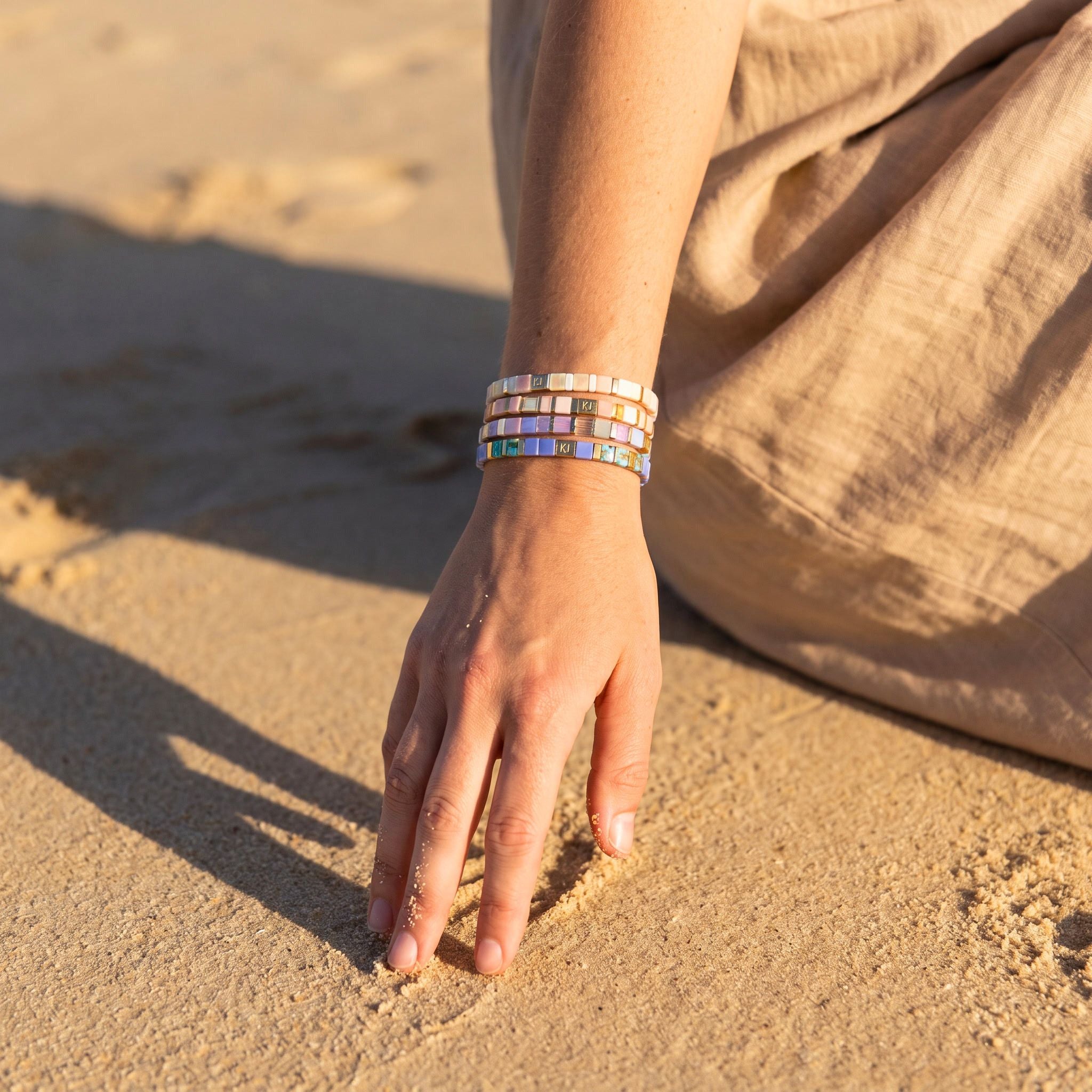 A close-up of a person's hand resting on sandy ground, wearing a stack of three colorful beaded bracelets. The person is dressed in a beige linen garment, and the background is a sandy beach under warm golden sunlight.