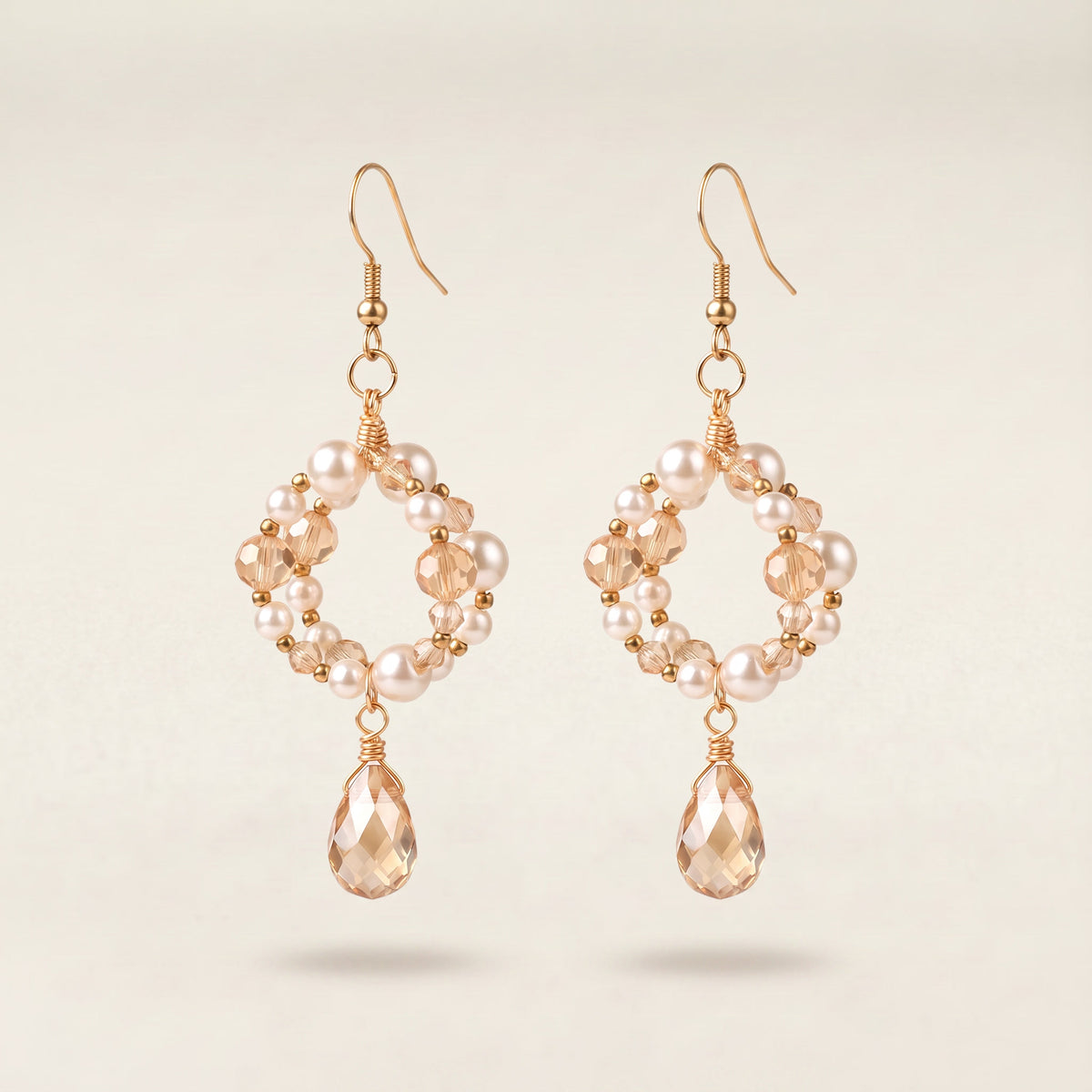 A pair of gold-tone dangle earrings featuring a circular arrangement of small white pearls and faceted peach-beige beads, with a faceted teardrop crystal hanging from each earring, set against a light beige background.