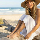 Woman in a white dress and straw hat sitting on driftwood by the ocean.