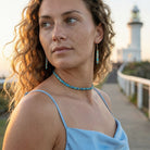 Woman with curly hair wearing a blue top and turquoise jewelry, standing in front of a lighthouse.