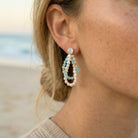 Close-up of a person wearing a pearl and gemstone earring with a blurred beach background