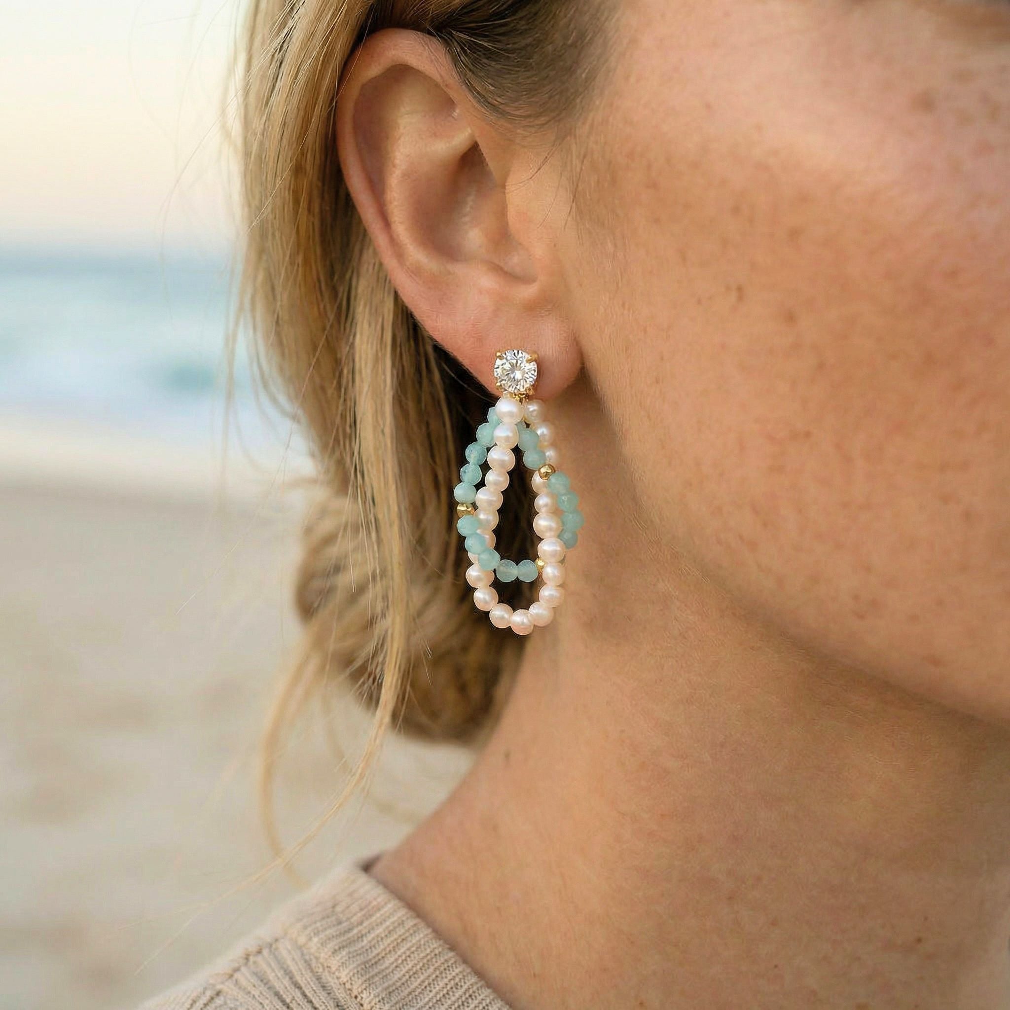 Close-up of a person wearing a pearl and gemstone earring with a blurred beach background