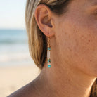 Close-up of a person wearing gold and turquoise earrings with a blurred beach background