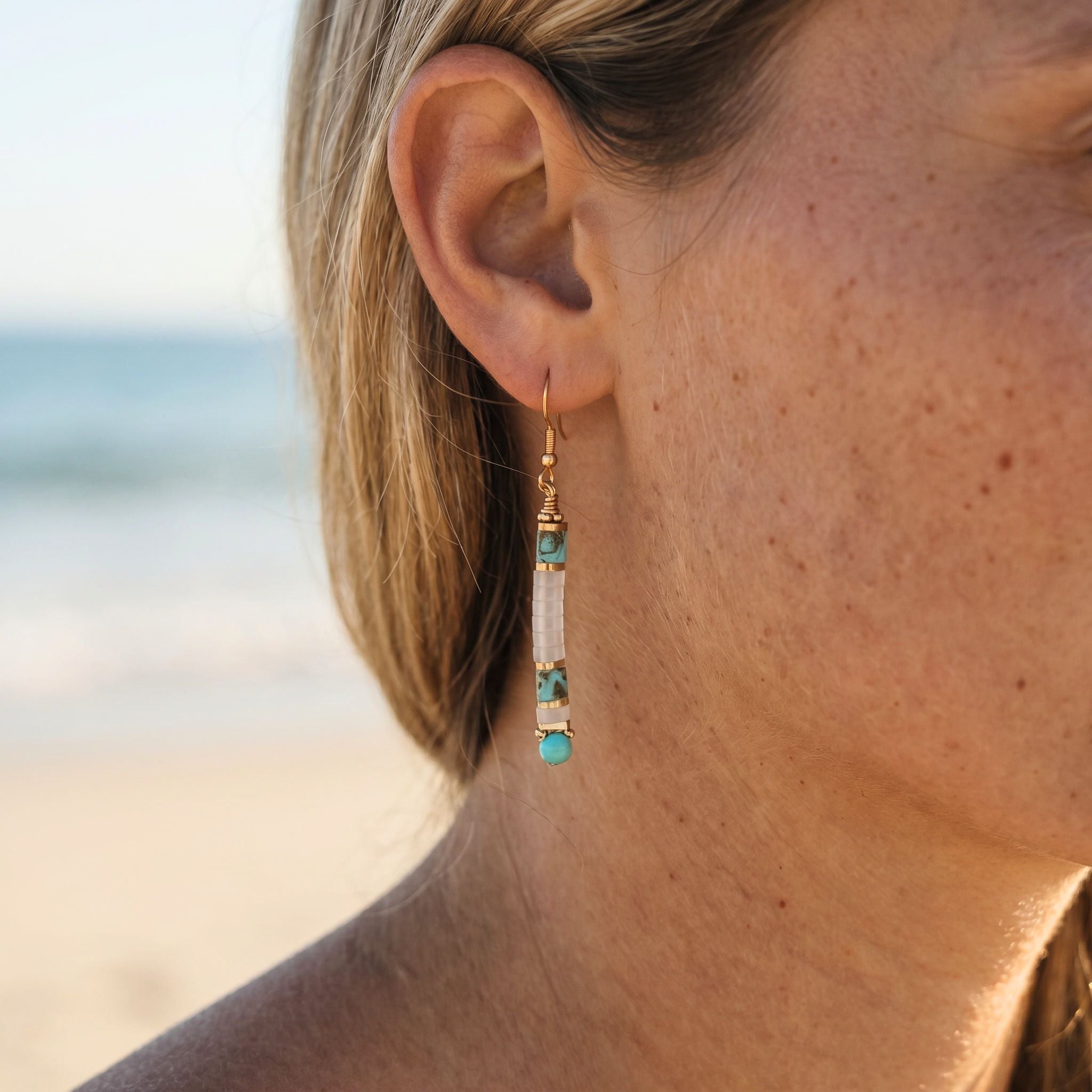 Close-up of a person wearing gold and turquoise earrings with a blurred beach background