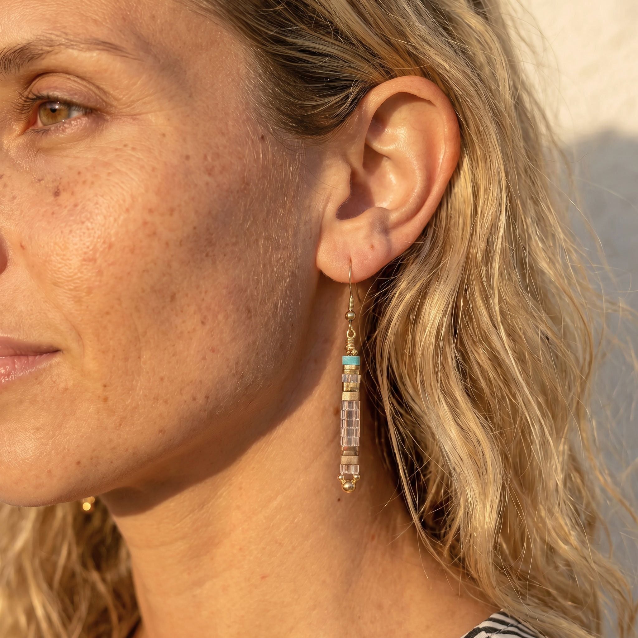 Close-up of a woman wearing gold earrings with a turquoise stone against a blurred natural background.
