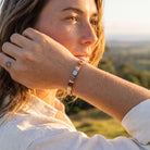 A close-up lifestyle shot of a woman wearing a slim metallic cuff bracelet with alternating rose gold and silver rectangular links, set against a blurred outdoor landscape background.