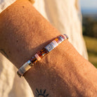 A close-up of a person's wrist wearing a beaded bracelet with amber, pink, white, and silver beads, featuring a central silver square engraved with 'KI'.