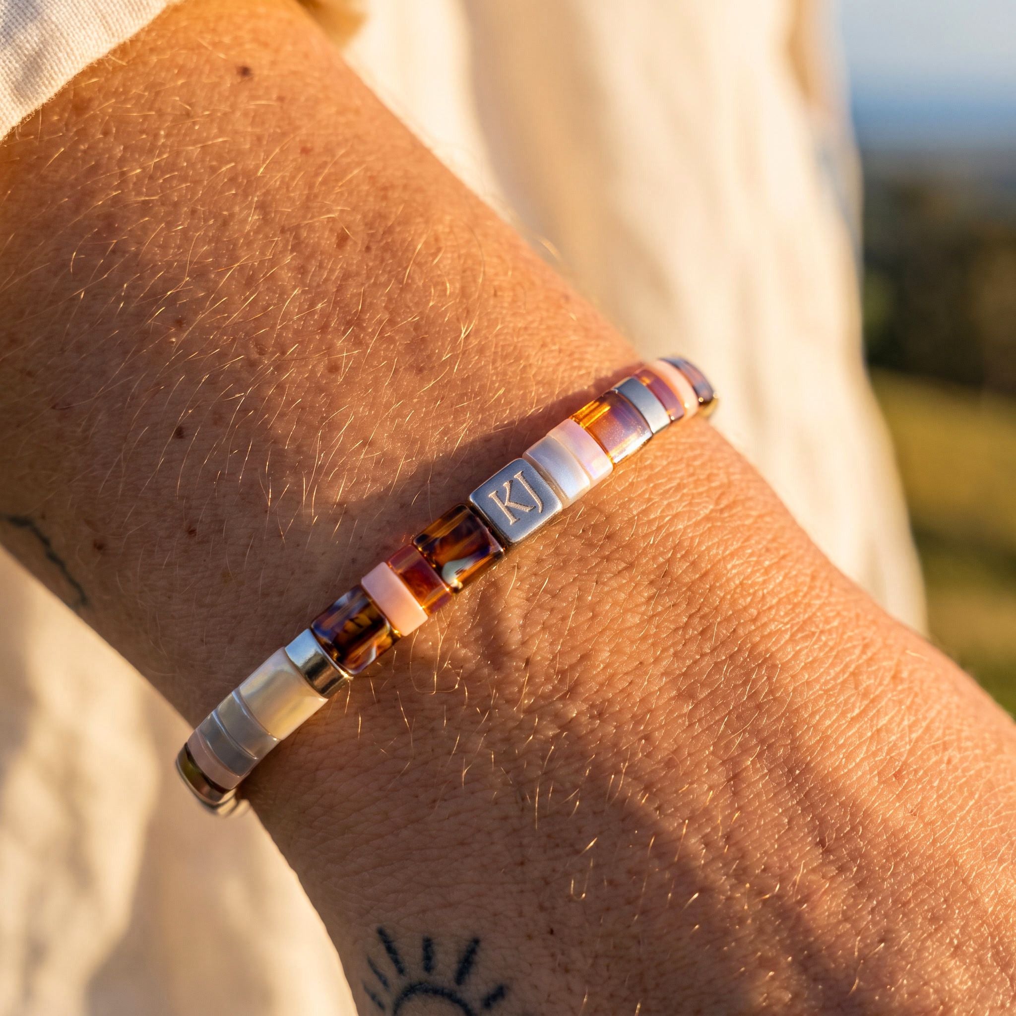 A close-up of a person's wrist wearing a beaded bracelet with amber, pink, white, and silver beads, featuring a central silver square engraved with 'KI'.