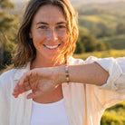 A smiling woman outdoors at sunset wearing a slim rectangular beaded bracelet on her wrist, with her arm raised to show it. She has freckles and light brown hair, wearing a white button-up shirt over a white ribbed top, with a blurred green hillside background.