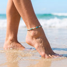 A close-up of a person's bare feet at the edge of the ocean, with a thin beaded anklet in turquoise, white, and orange beads worn on the ankle.