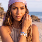 A woman on a beach wearing a lavender headscarf and a multi-strand beaded bracelet with iridescent blue, purple, and white beads.