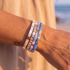 A close-up of a person's wrist wearing five stacked beaded bracelets in pastel colors, photographed outdoors with a blurred beach and ocean background.