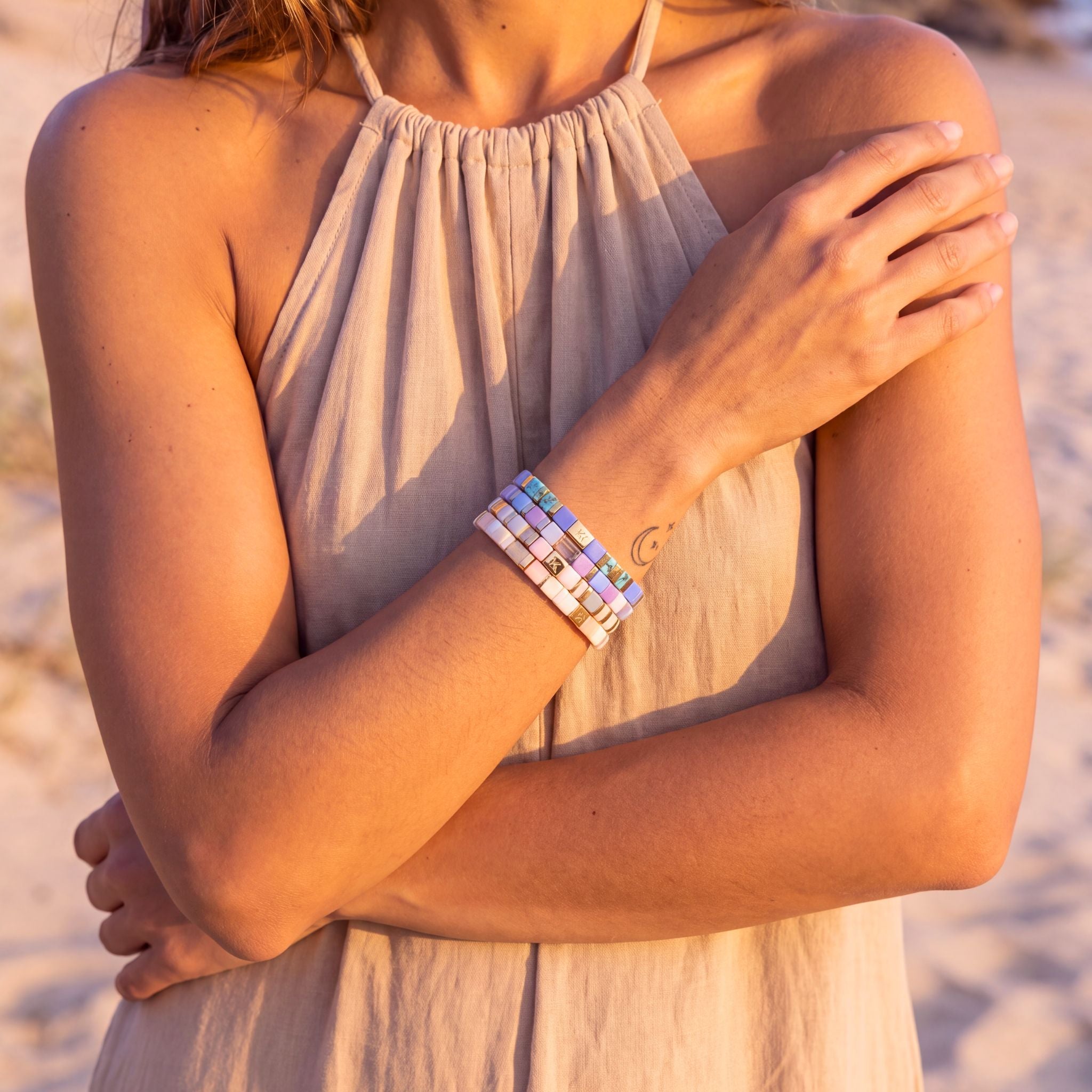 A woman outdoors at sunset wearing a stack of three colorful beaded bracelets on her wrist, with her arms crossed over her chest.