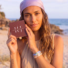 A woman on a beach holds a square brown box labeled 'KAWAI JEWELLERY' while wearing colourful beaded bracelets on her wrist. The background shows sand, rocks, and the ocean.