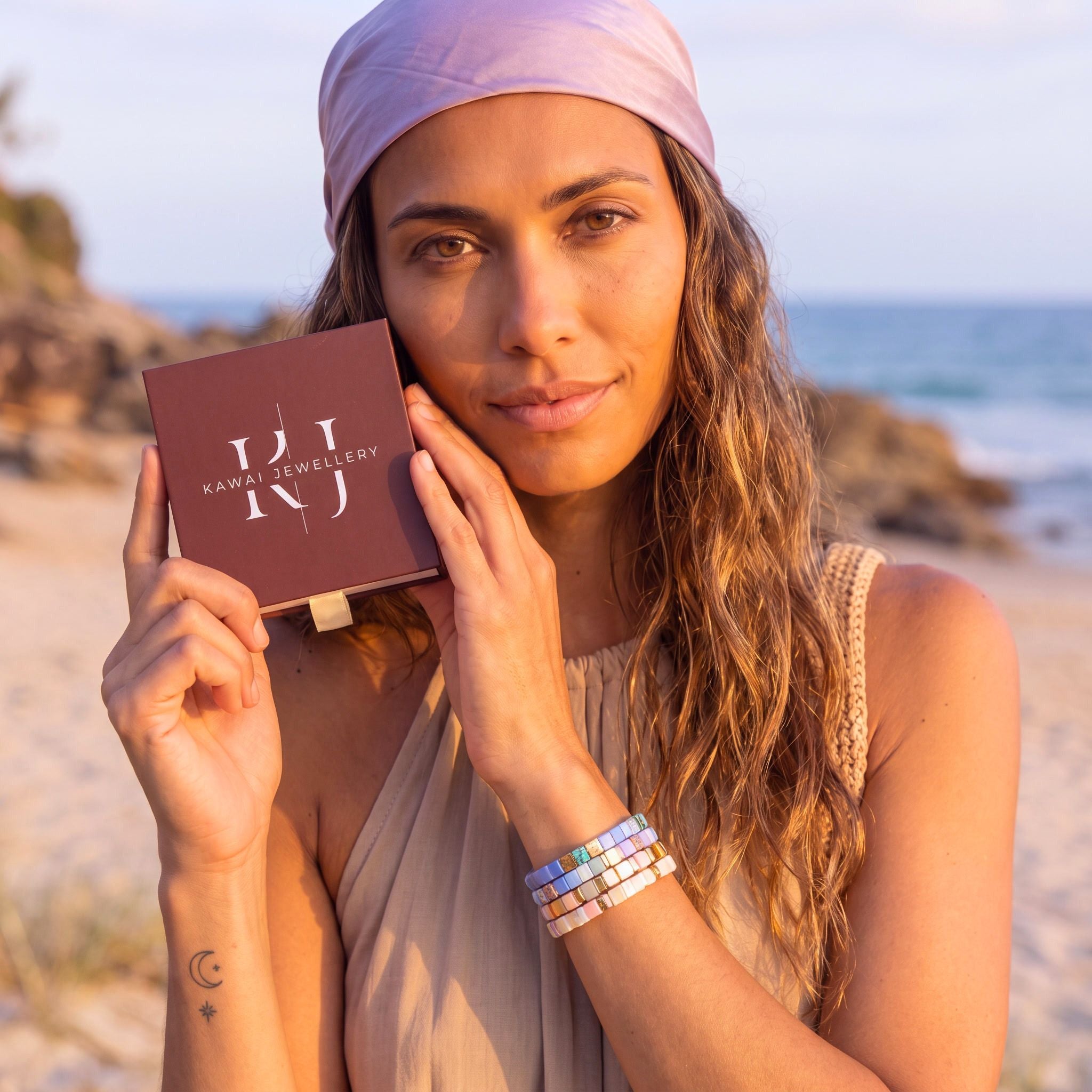 A woman on a beach holds a square brown box labeled 'KAWAI JEWELLERY' while wearing colourful beaded bracelets on her wrist. The background shows sand, rocks, and the ocean.