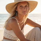 Woman sitting on a beach wearing a straw hat and crochet top.