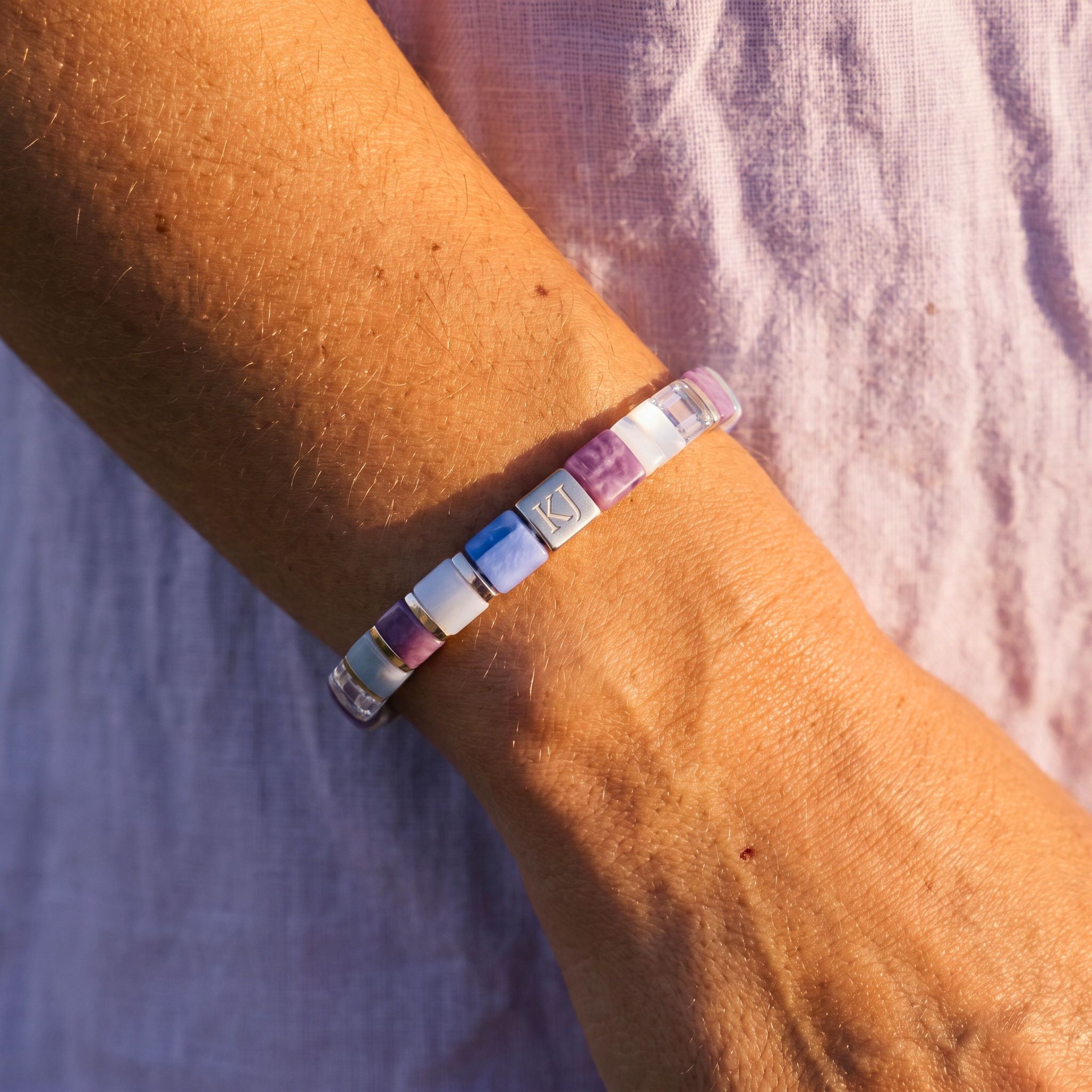 A close-up of a person's wrist wearing a beaded stretch bracelet made of alternating translucent square beads in purple, blue, white, and pink, with one silver-toned bead featuring a white crescent moon symbol.