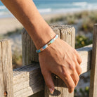 Hand wearing a colorful beaded bracelet on a wooden railing with a beach in the background