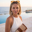 A woman with blonde hair wearing amber-framed sunglasses on her head, a white sleeveless top, and pearl drop earrings, holding a woven straw clutch with brown trim. She is photographed outdoors by a pool with the ocean in the background.