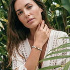 Woman wearing a white lace top and bracelet with greenery in the background