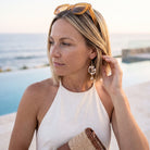 A woman wearing a white halter top and dangling pearl-like earrings stands outdoors near a pool and ocean, holding a woven clutch. Sunglasses rest on her head.