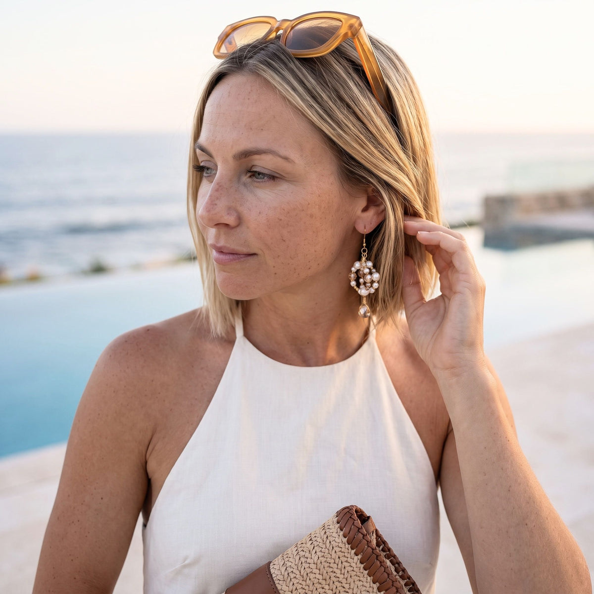 A woman wearing a white halter top and dangling pearl-like earrings stands outdoors near a pool and ocean, holding a woven clutch. Sunglasses rest on her head.