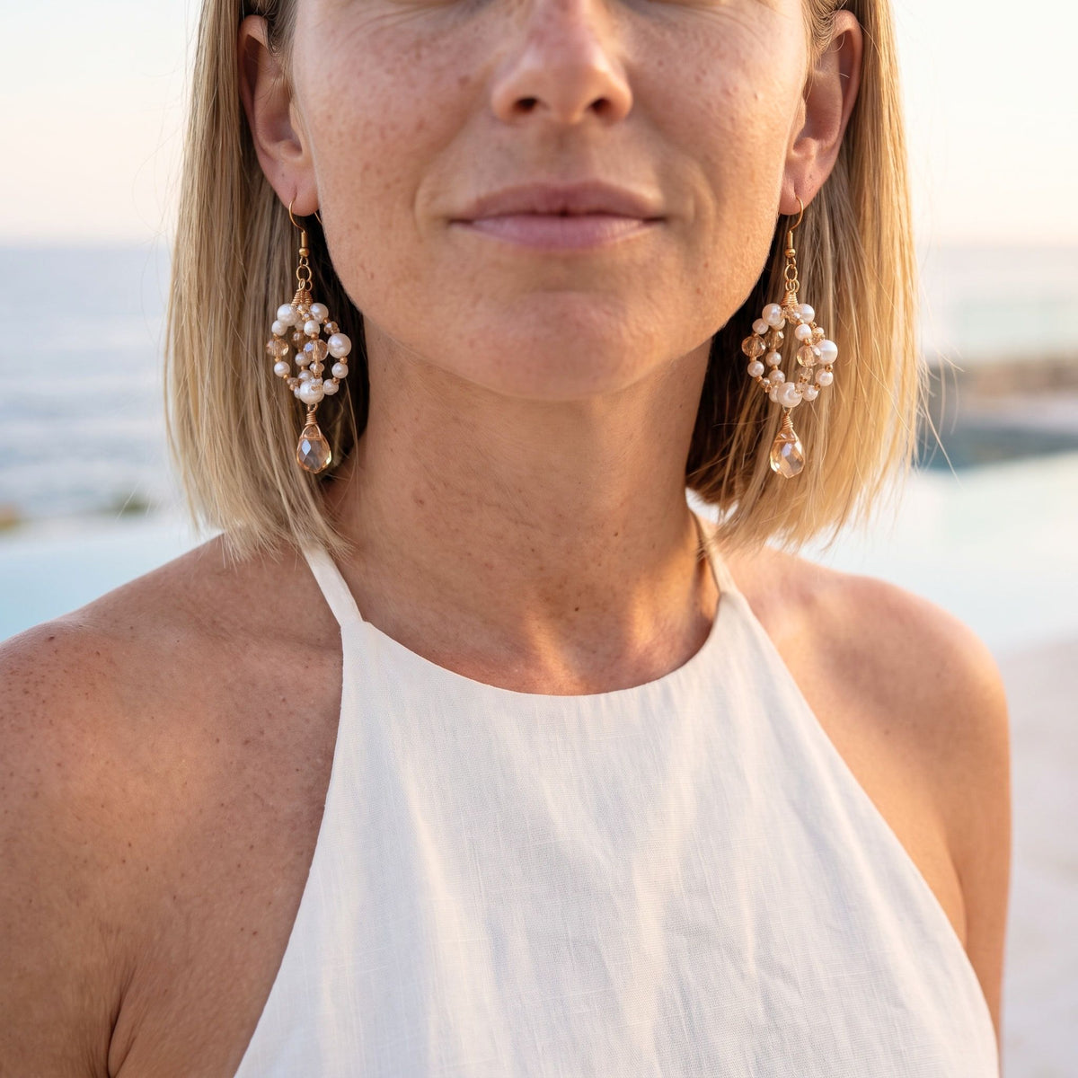 A close-up of a woman wearing a pair of gold-toned drop earrings with clusters of white pearl-like beads and teardrop charms, photographed outdoors near a pool or water feature.