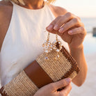 A close-up lifestyle shot of a woman holding a woven straw clutch bag with brown leather trim, while wearing dangling gold hoop earrings adorned with white pearls and clear crystal drops.