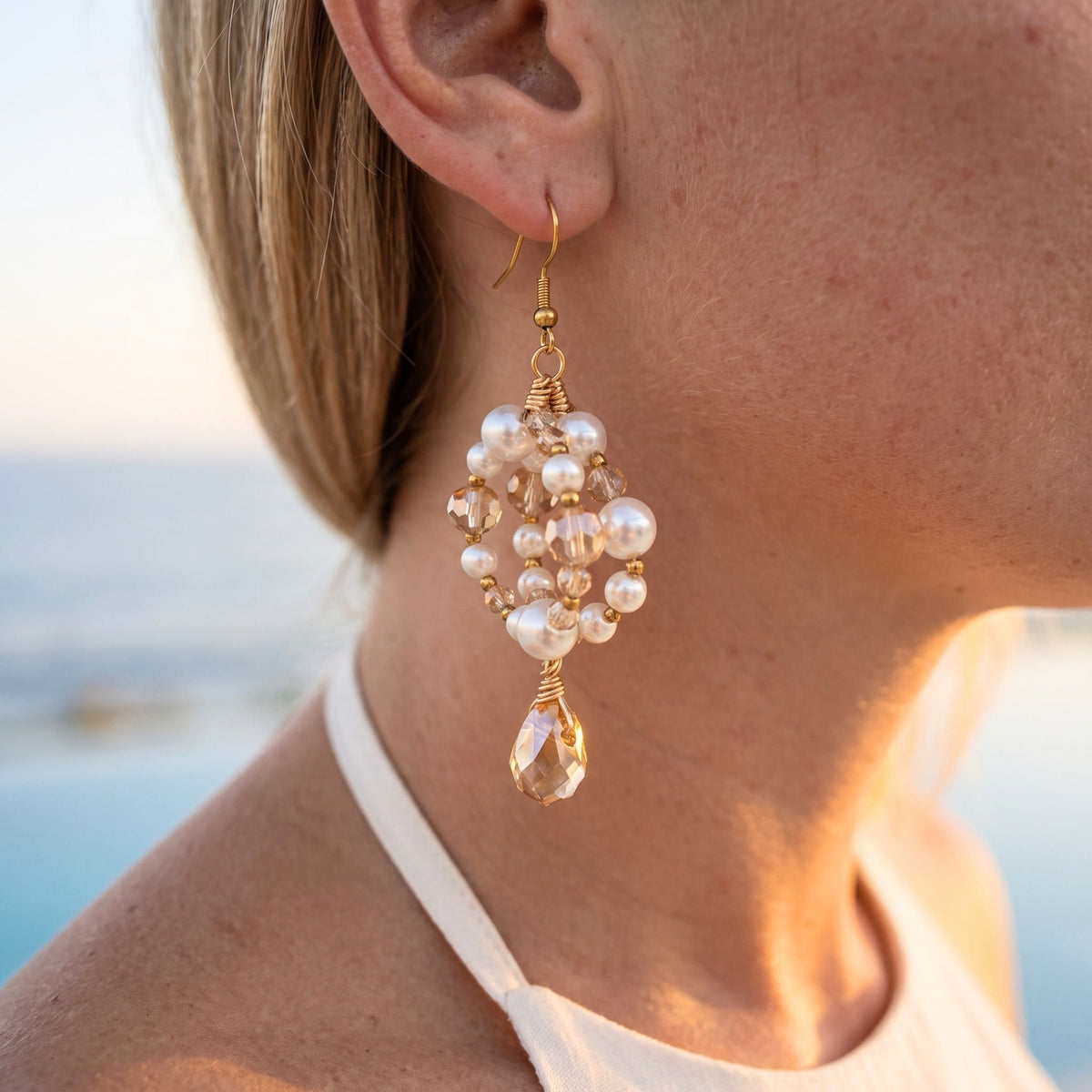 A close-up of a woman's ear and neck wearing a dangling pearl earring with gold-tone metal and a faceted clear teardrop charm, set against a softly blurred outdoor background.