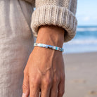 A close-up of a person's wrist wearing a silver-toned beaded bracelet with light blue, white, and dark beads, set against a blurred beach background.