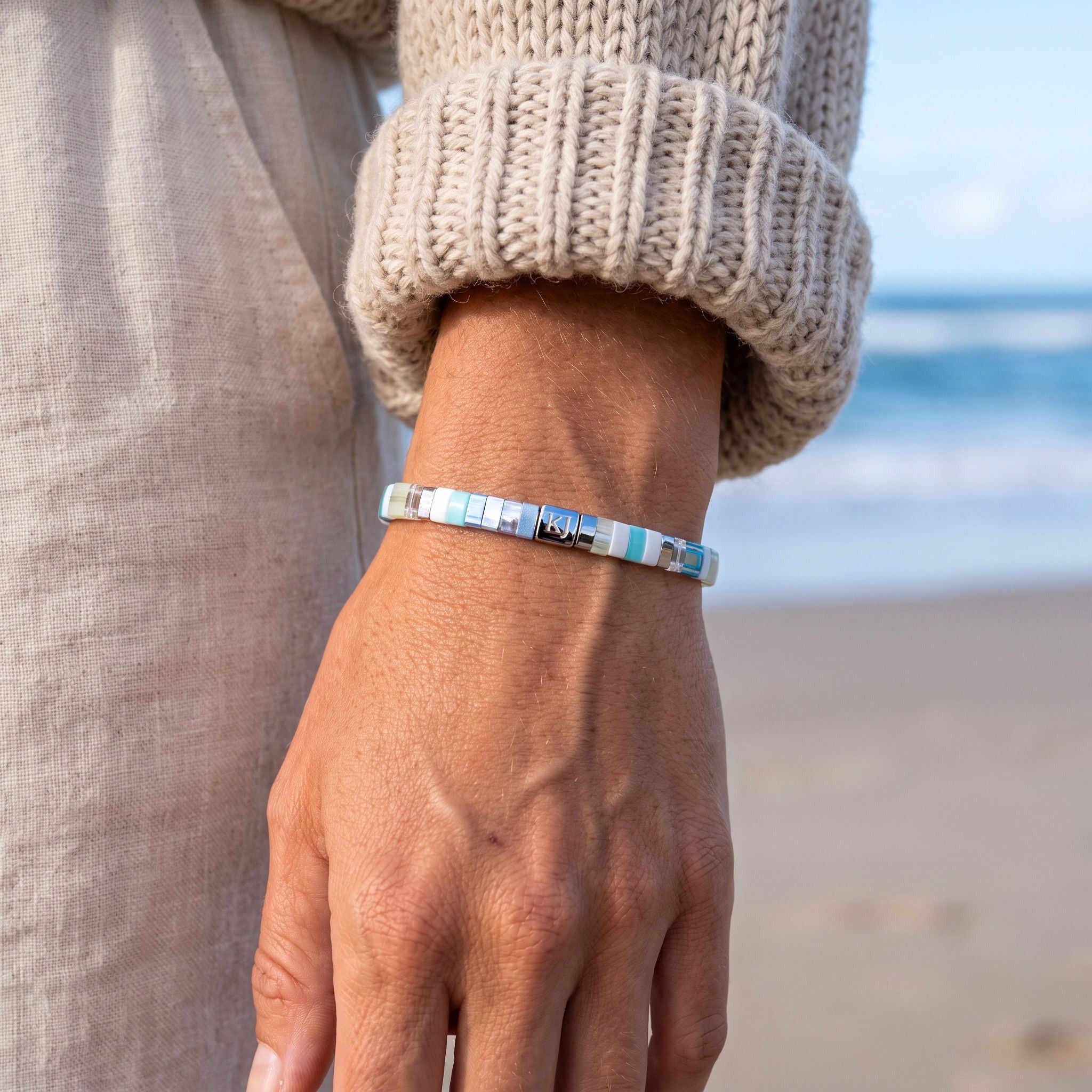 A close-up of a person's wrist wearing a silver-toned beaded bracelet with light blue, white, and dark beads, set against a blurred beach background.