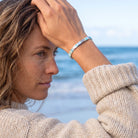 A woman in a beige knit sweater wearing a silver-toned beaded bracelet, posing with her hand on her head against a blurred beach and ocean background.