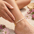 Close-up of a person's ankle with a bracelet, surrounded by pink flowers on a natural surface.