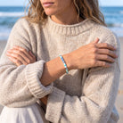 A woman wearing a chunky knit beige sweater stands on a beach, showcasing a light blue beaded bracelet on her wrist.