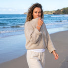 A woman stands on a sandy beach wearing a chunky knit beige sweater over a white top and light beige pants, with the ocean and distant cliffs in the background.