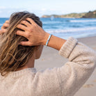 A woman on a beach wearing a silver bracelet with a blue and white patterned stone inlay, seen from behind as she looks out toward the ocean.