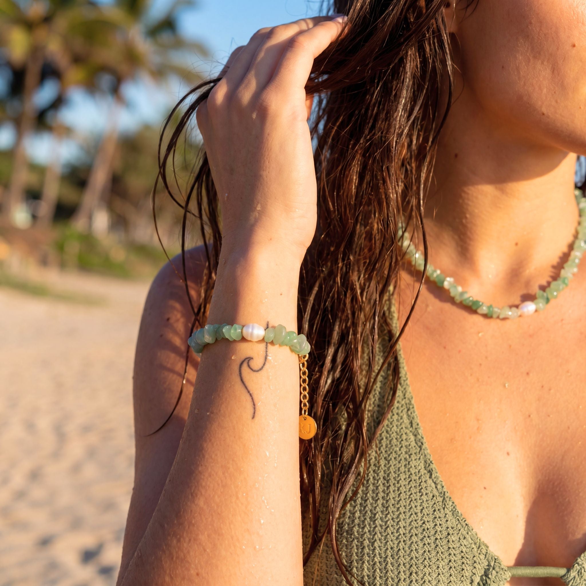 Woman on a beach wearing a green beaded bracelet and necklace, with blurred palm trees in the background.