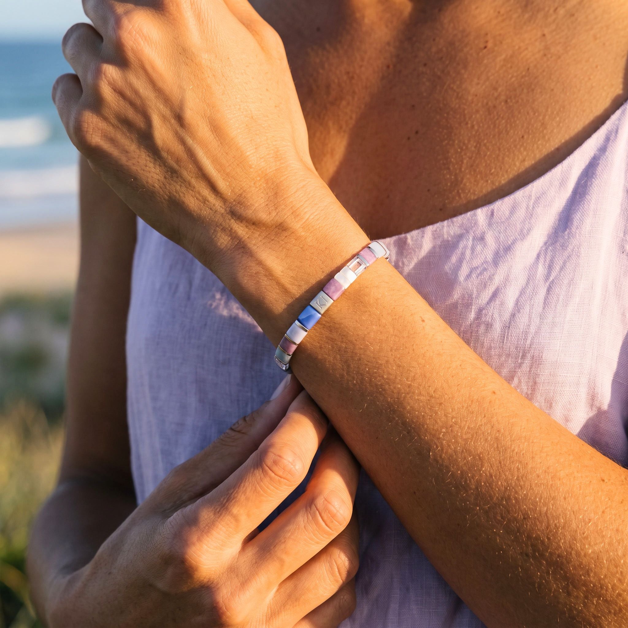 A close-up lifestyle shot of a person outdoors by the beach, wearing a thin beaded bracelet with pastel pink, blue, and white beads on their wrist.