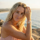 Woman with blonde hair and freckles sitting on a beach, holding her hand to her face.