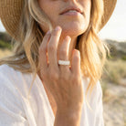Woman wearing a straw hat and white top with a close-up of her hand showing a ring.