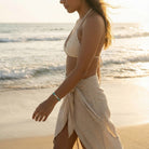 Woman in a white bikini top and beige wrap on a beach at sunset.
