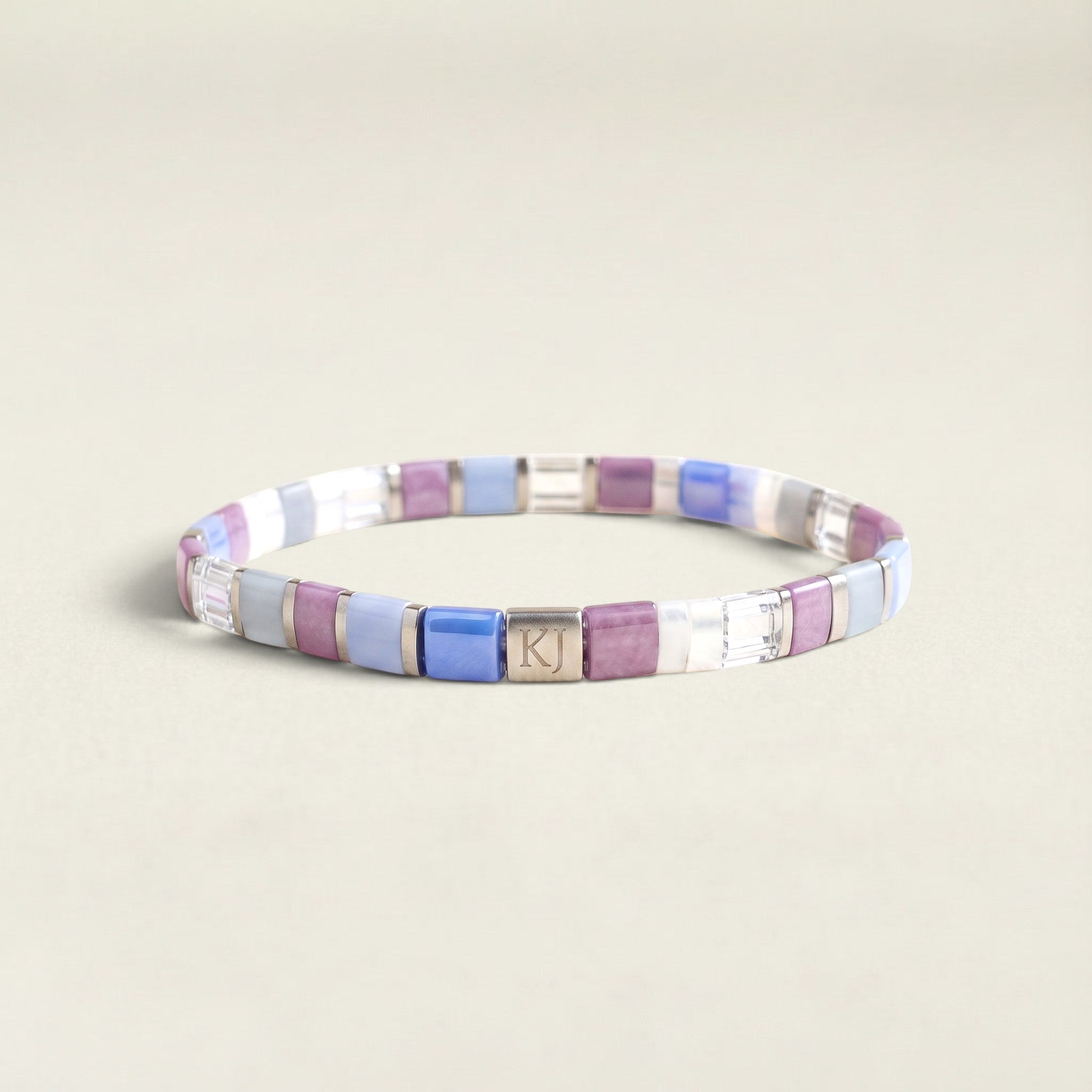 A personalized beaded stretch bracelet with square tiles of lavender, light blue, white, and metallic silver, featuring a central silver charm engraved with the initials 'KJ'.