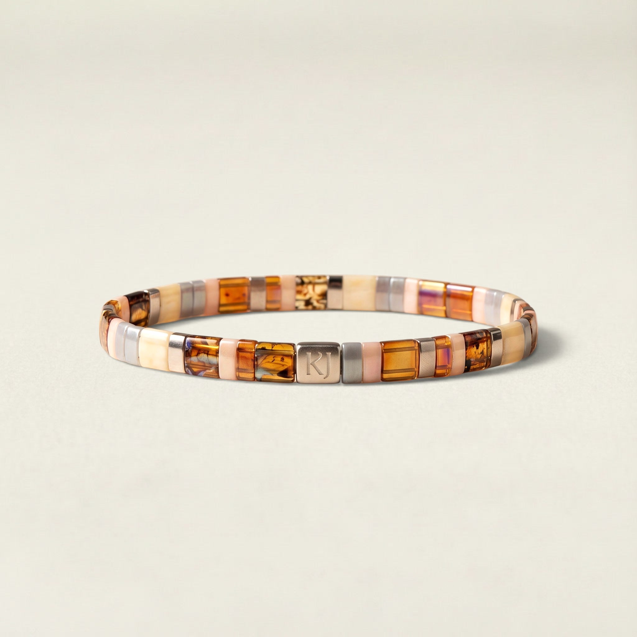 A beaded stretch bracelet featuring rectangular amber, brown, and cream-colored stones, accented by a silver-toned metal square with an engraved letter 'N', set against a light beige background.
