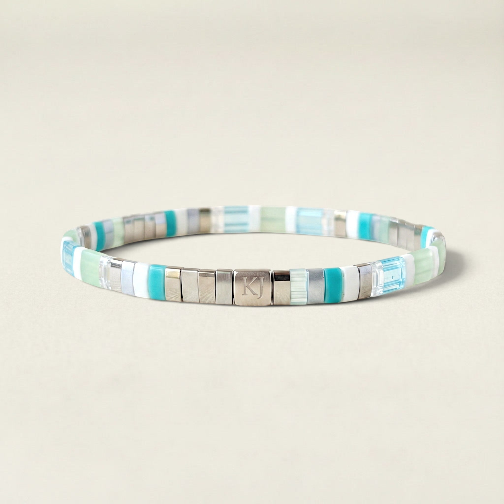 A beaded stretch bracelet featuring alternating turquoise, mint green, light blue, and silver metallic square beads, with a central silver clasp engraved with the initials 'RJ'.