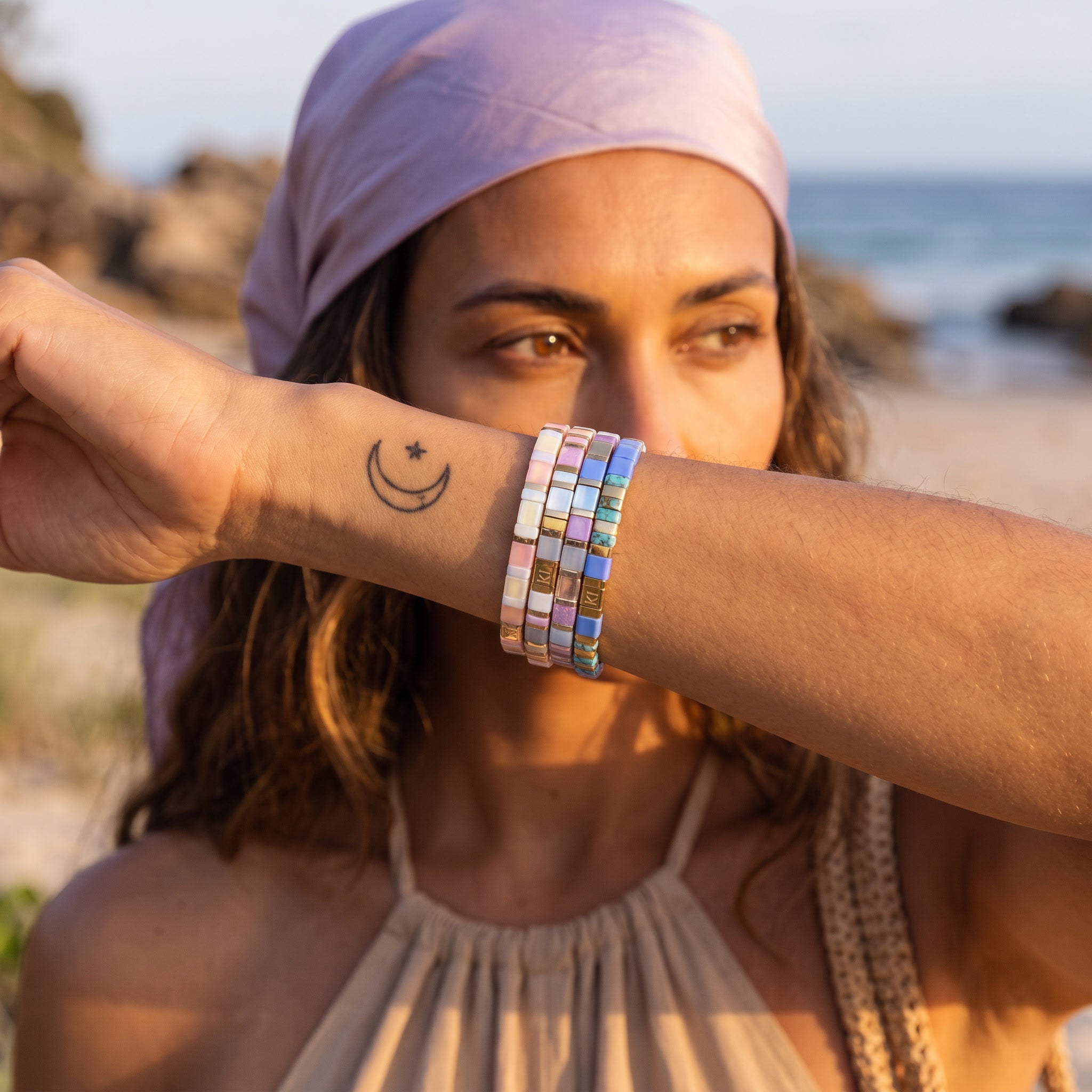 A woman at the beach wearing a stack of colorful beaded bracelets on her wrist, with a lavender headscarf and beige halter top in the background.