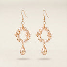 A pair of gold-tone dangle earrings featuring a circular arrangement of small white pearls and faceted peach-beige beads, with a faceted teardrop crystal hanging from each earring, set against a light beige background.
